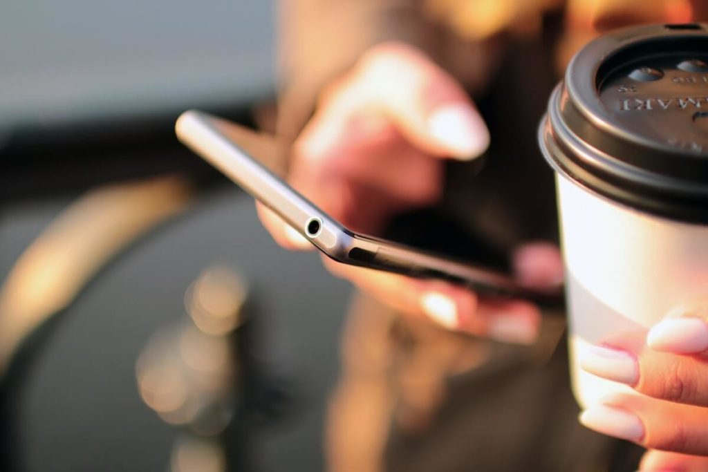 Henkilö selaa älypuhelinta take away kahvi toisessa kädessään
