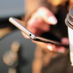 Henkilö selaa älypuhelinta take away kahvi toisessa kädessään
