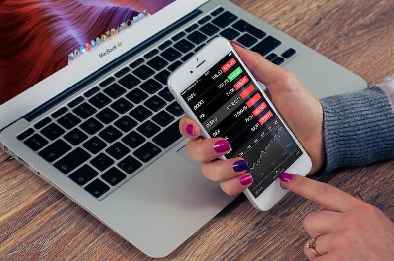 Sijoittamisen alkeita opetteleva nainen kädessään iPhone, jonka näytöllä näkyy osakemarkkinoiden numerot ja kaaviot